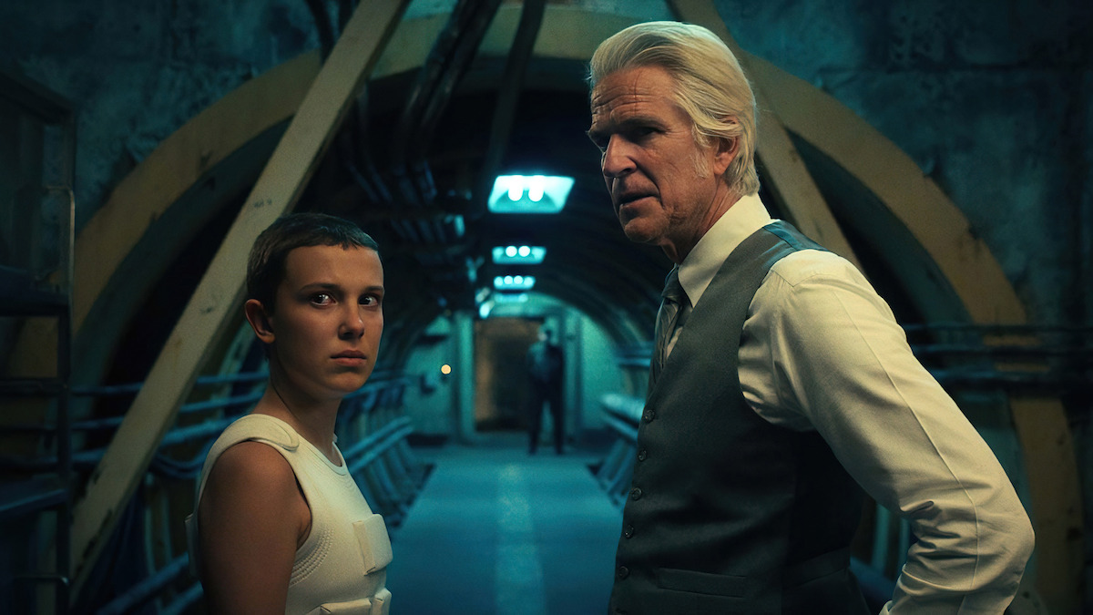 A serious young girl and an older man stand in a dimly lit industrial tunnel with pipes on the walls, creating a tense and mysterious atmosphere.