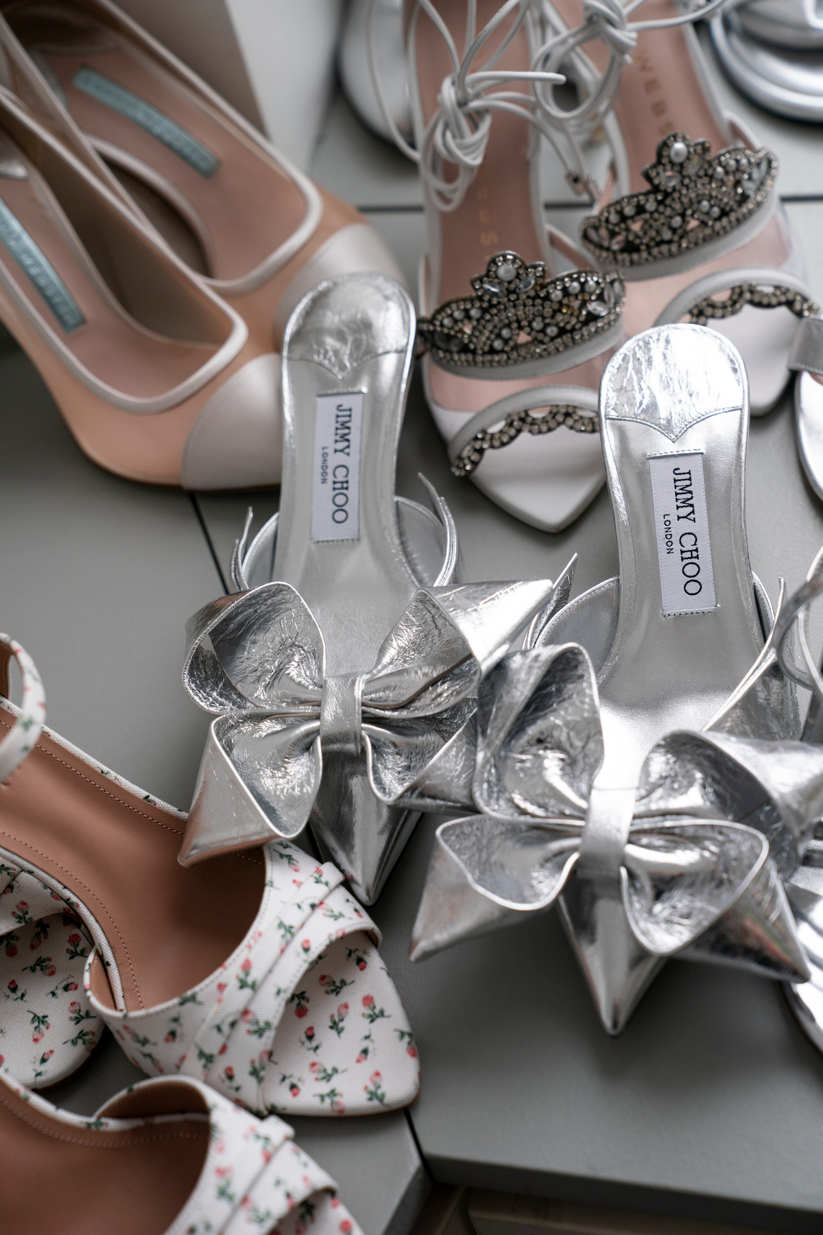 Collection of shoes including silver Jimmy Choo bow heels on a gray surface.