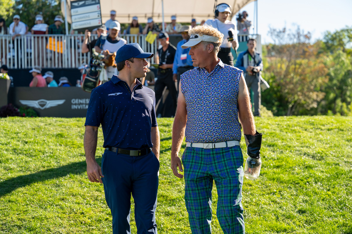 Two men, one in a navy golf outfit and the other in a sleeveless patterned shirt and plaid pants, stand on a golf course talking during a sunny day, with spectators and a tournament sign visible in the background.