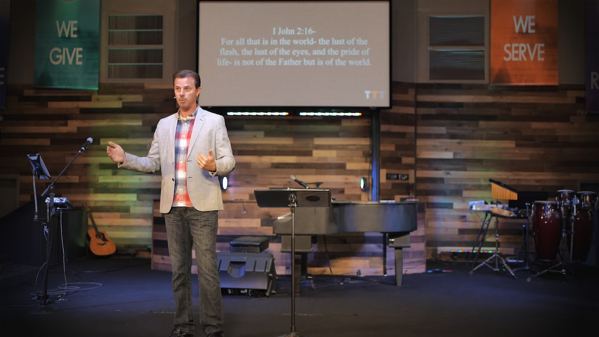 A man speaks on stage in a church with wooden walls, musical instruments, colorful banners reading "WE GIVE" and "WE SERVE," and a large screen displaying a Bible verse from 1 John 2:16 behind him.