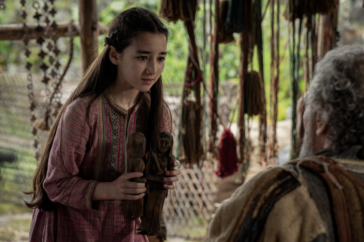 A young girl in traditional clothing holds two wooden figurines while speaking to an older person in a rustic, handcrafted setting, creating a thoughtful and intimate mood.