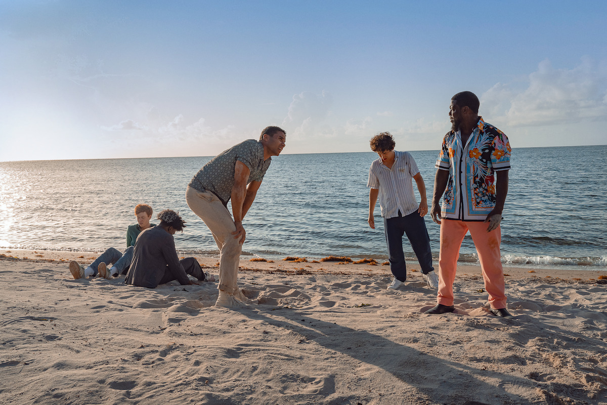 Five people on a sandy beach by the ocean, some standing and talking while others sit near the shoreline, under a clear blue sky in the late afternoon.