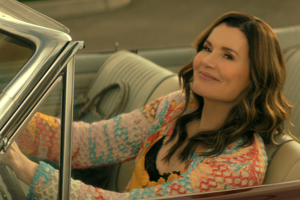 Geena Davis as Renee, with long hair wearing a colorful sweater, sitting in the driver's seat of a vintage convertible car, outdoors in daylight, looking to the side while driving.
