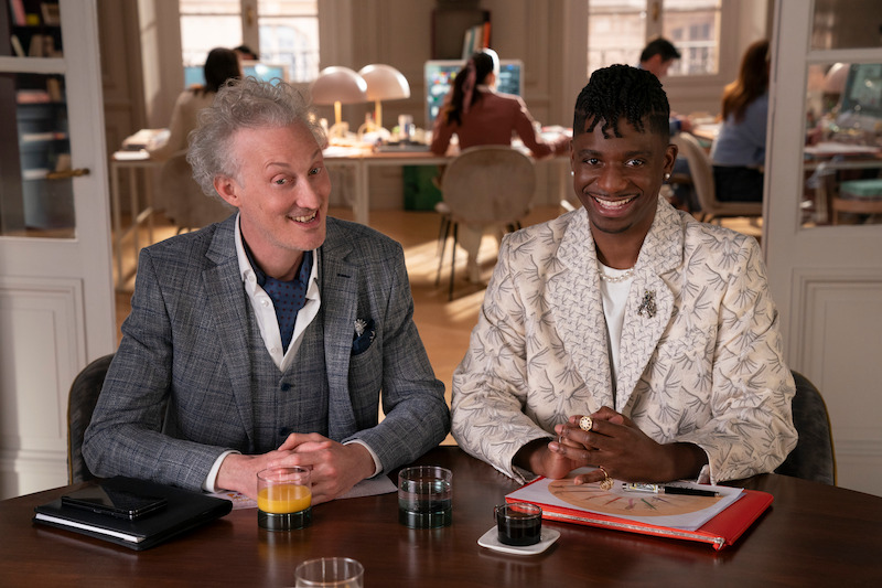  Bruno Gouery as Luc and Samuel Arnold as Julie sit in an office together smiling in Season 4 of 'Emily in Paris'