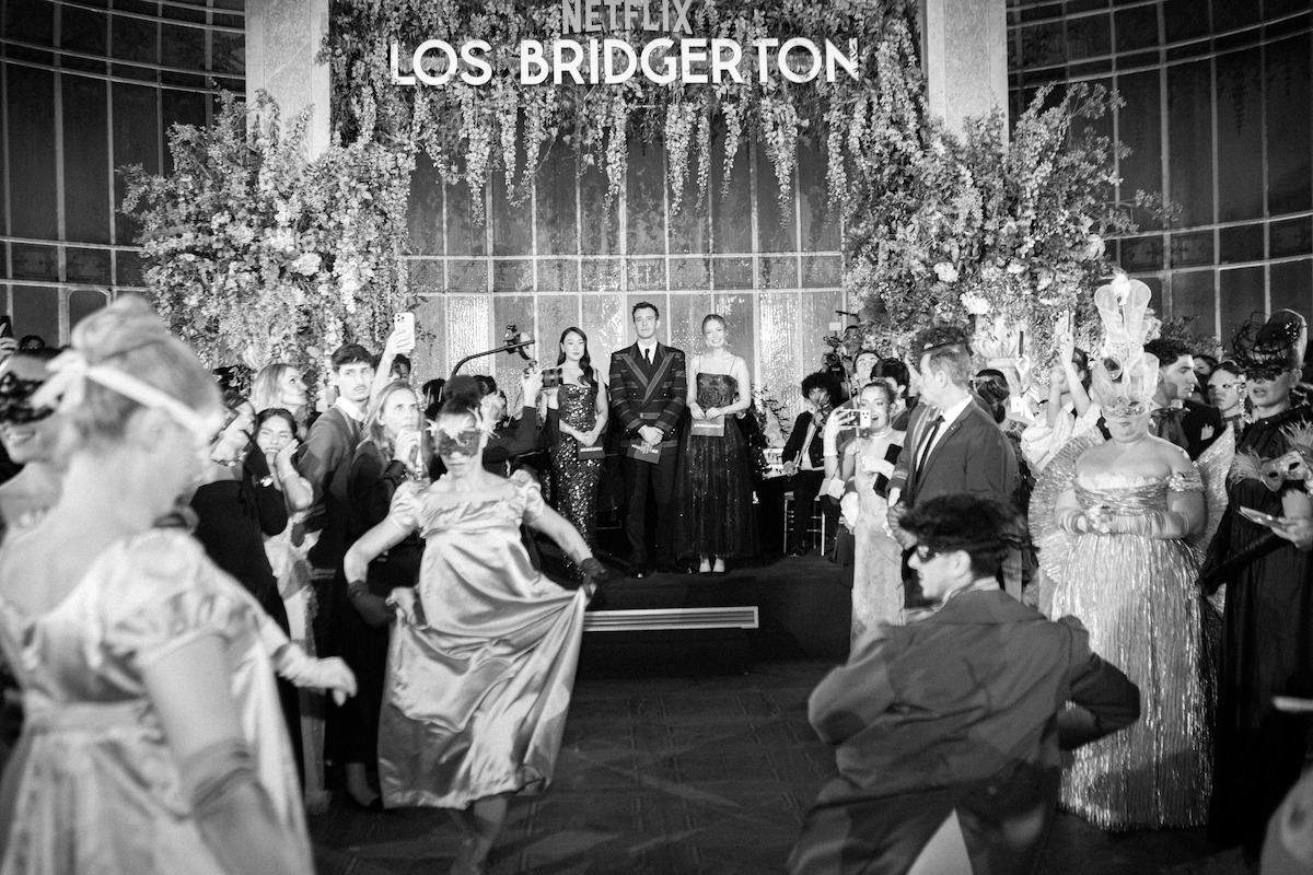 Um grande salão de baile, com pessoas usando trajes de época. Elas dançam e interagem. Logo acima delas está uma placa com os dizeres “Netflix Los Bridgerton”, com decorações ornamentadas e um ambiente formal.