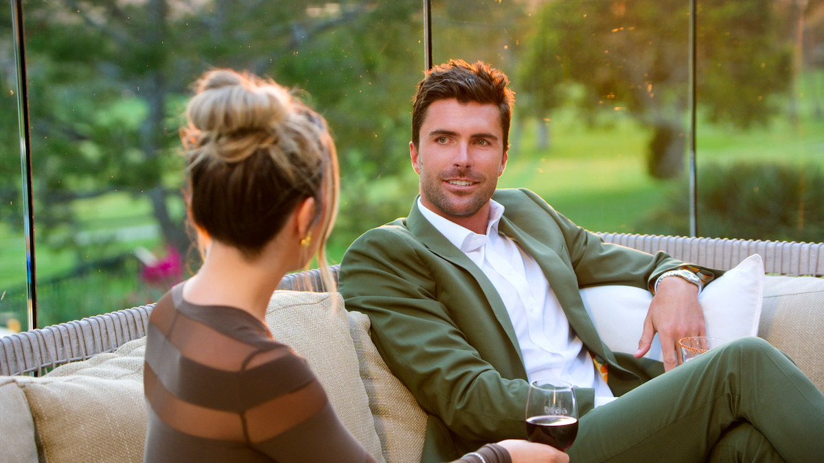 A man in a green suit and a woman sitting on an outdoor couch have a relaxed conversation with wine, surrounded by greenery and glass panels in a serene, warm sunset setting.