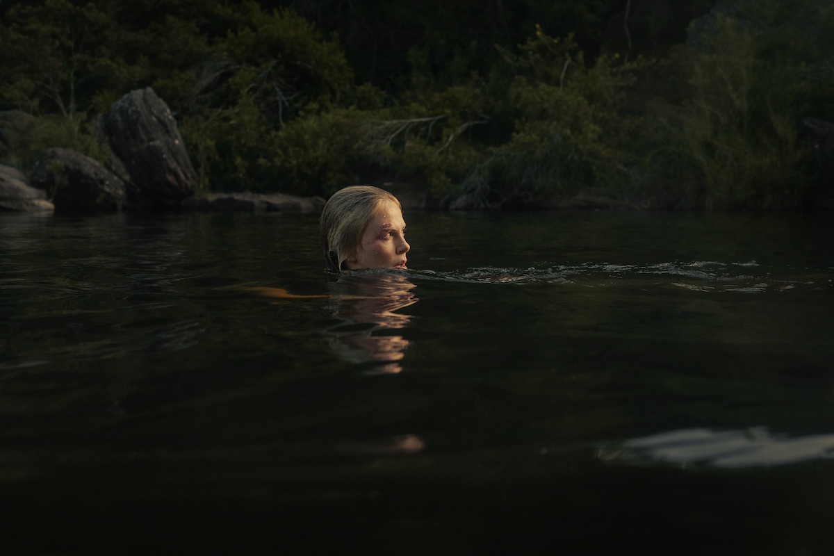 A person swims in a calm, dark natural body of water surrounded by dense green foliage and large rocks, partially submerged with only their head showing above the water near a forested shoreline at dusk.