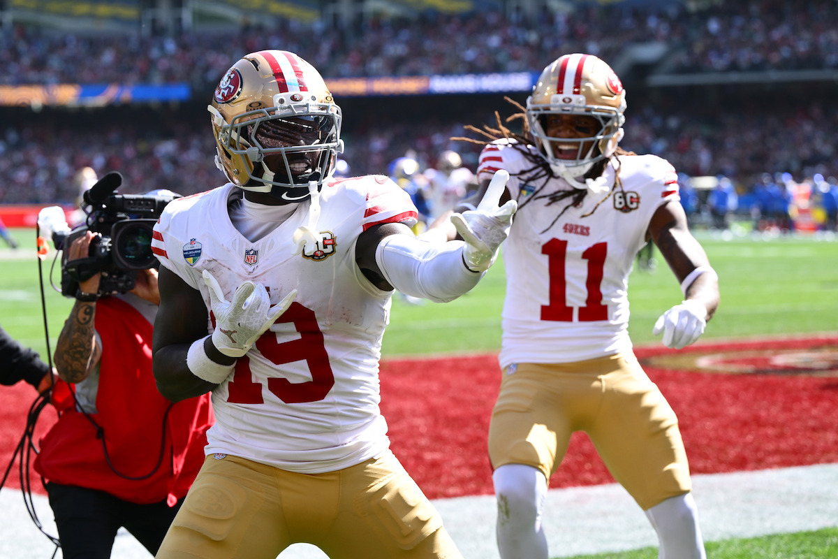 Two San Francisco 49ers football players celebrate in the end zone during a daytime game, with a crowded stadium and a cameraman capturing the scene.
