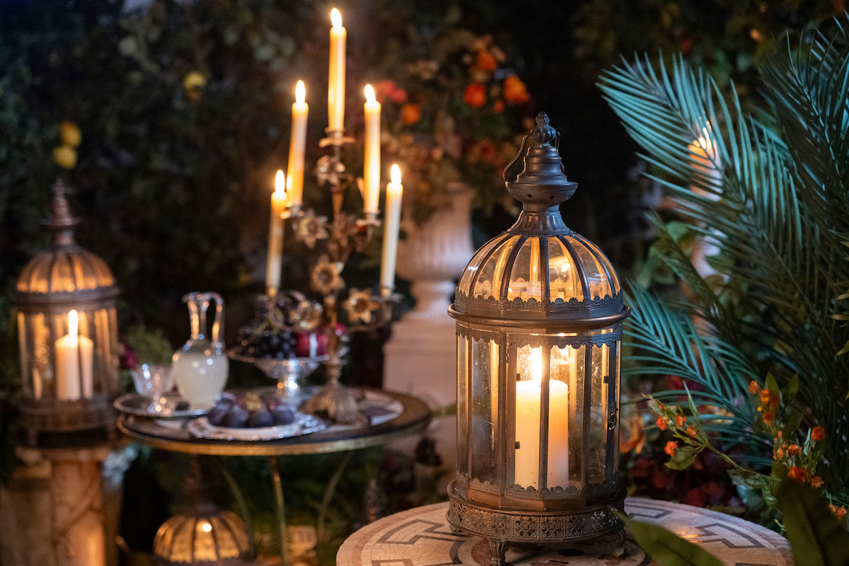 Antique lanterns with lit candles on ornate tables, surrounded by greenery and flowers, with a candelabrum, fruit, and a pitcher, creating a warm, romantic, outdoor evening setting.