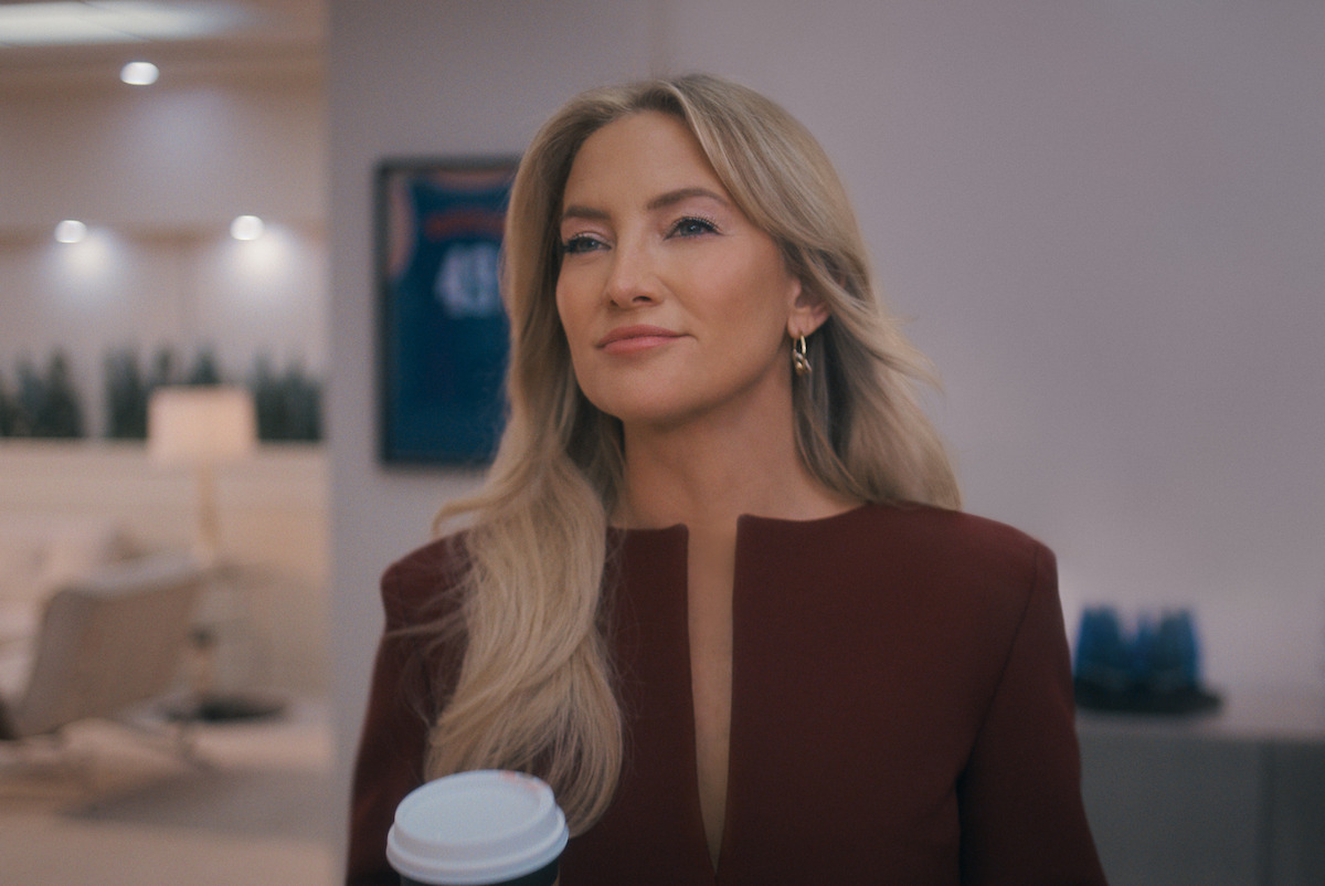 Woman with long blonde hair in a maroon outfit holding a coffee cup, standing in a modern, well-lit office with minimalist decor and a framed sports jersey on the wall in the background.