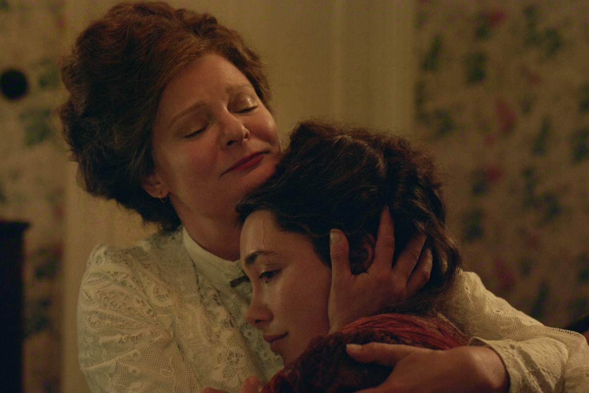 Two women in vintage clothing share an emotional embrace in a warmly lit, floral-wallpapered room, suggesting comfort and support in a historical or period setting.