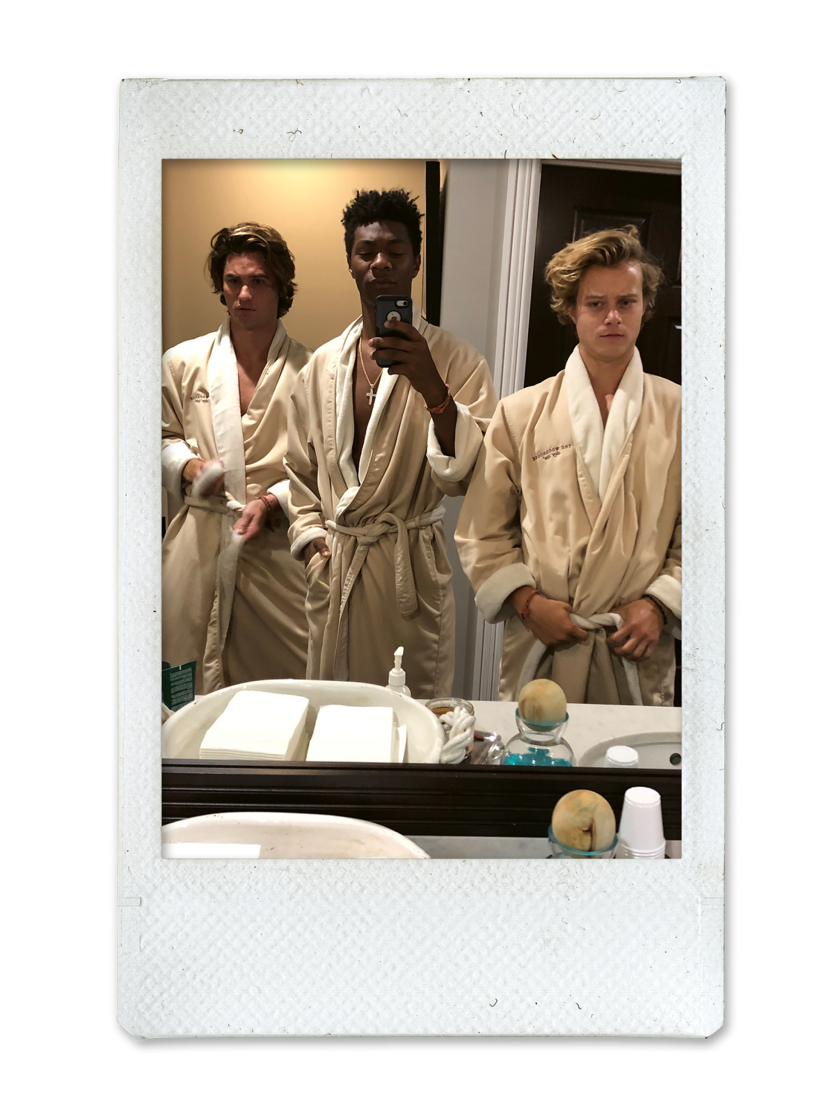 Three young men in beige bathrobes stand together in a bathroom, posing for a mirror selfie. The counter holds toiletries, towels, and soaps. The scene is casual and relaxed, set in what appears to be a hotel or spa bathroom.