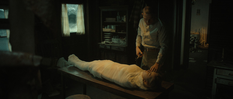 A person wrapped entirely in white bandages lies on a table while another person stands beside them holding scissors in a dimly lit room.