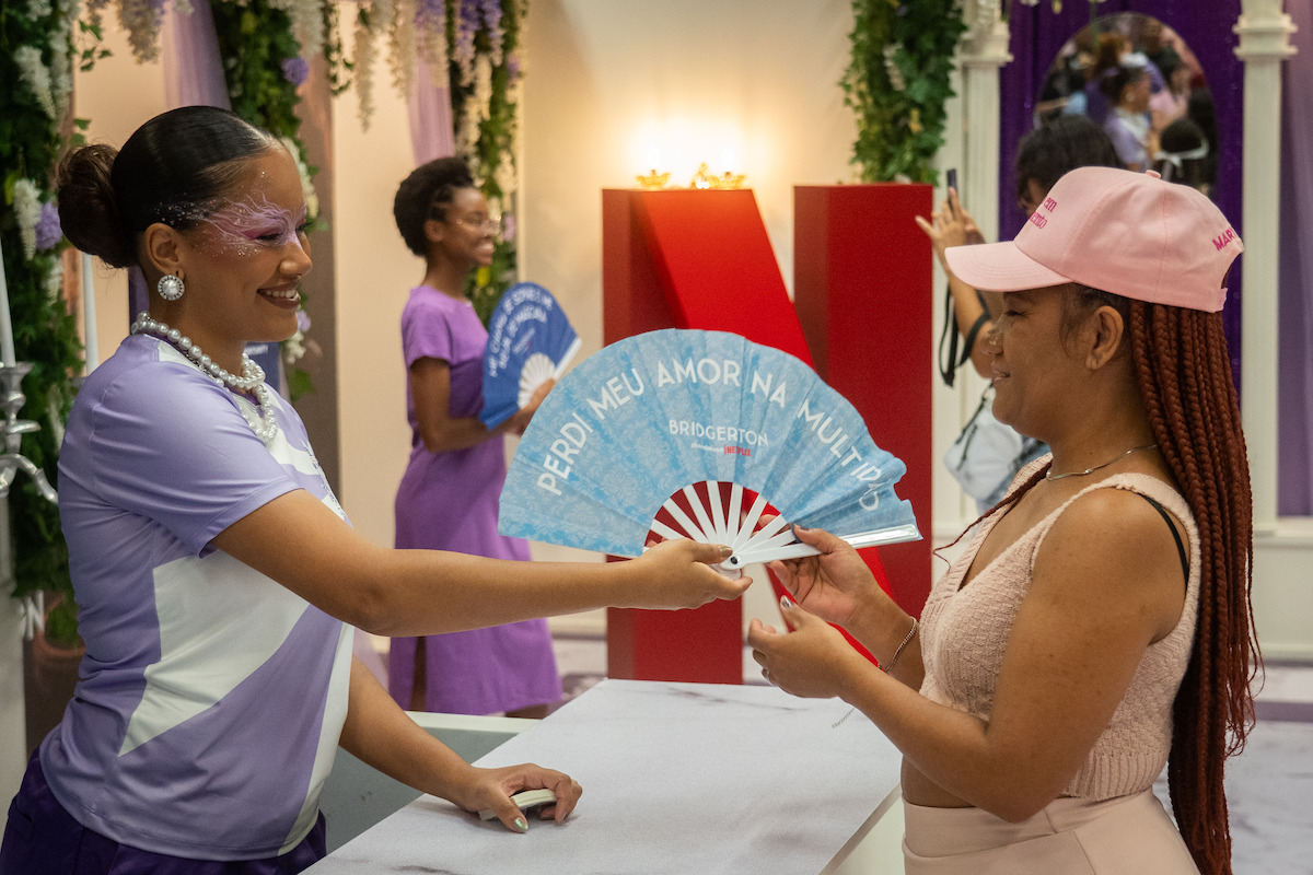 Duas mulheres sorridentes interagem em um evento realizado em ambiente interno, com plantas decorativas e um grande N vermelho ao fundo. Por cima de um balcão, uma delas entrega um leque azul à outra.