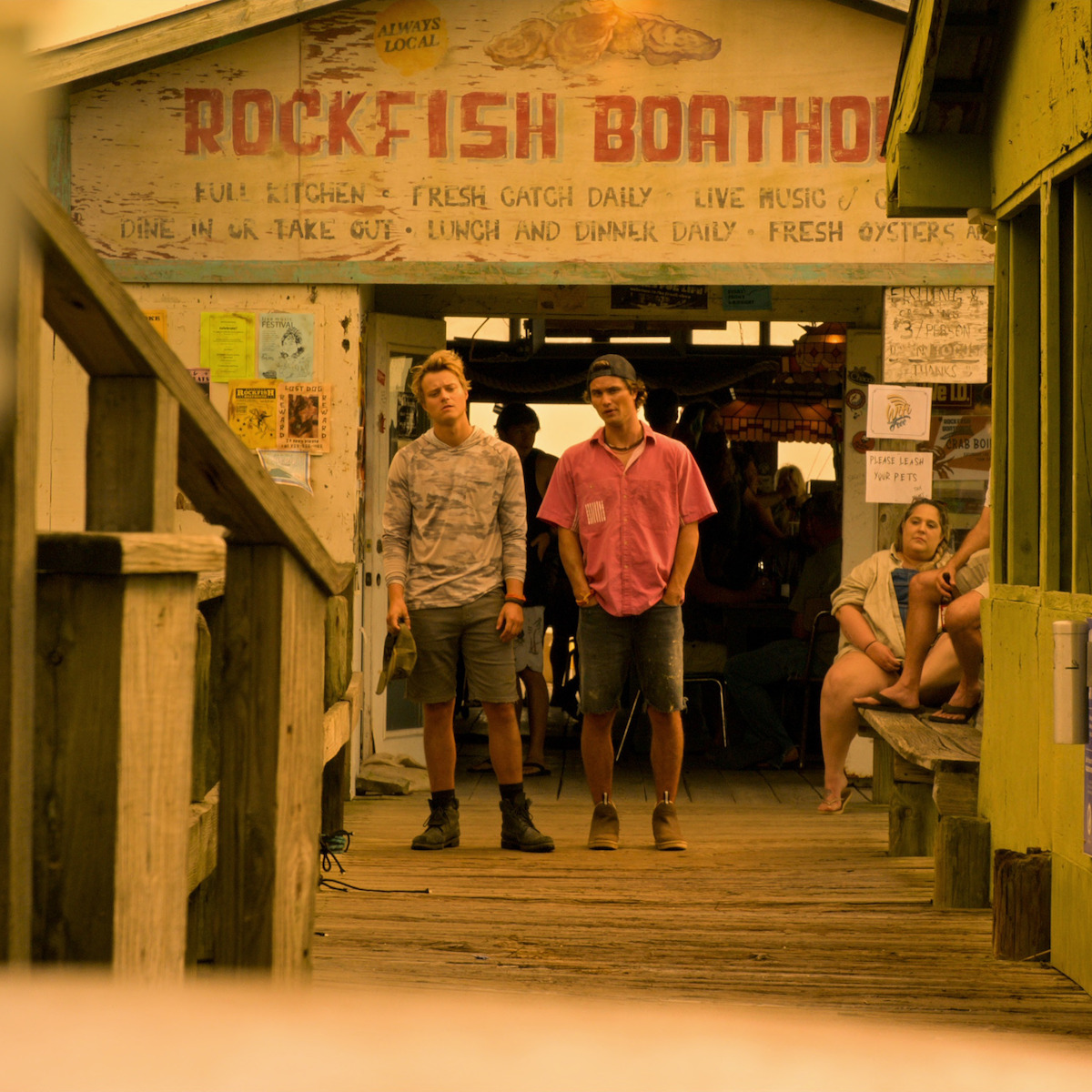 Dois homens jovens estão em pé em um píer de madeira em frente ao Rockfish, cercados de placas e pessoas sentadas em bancos. É um ambiente de restaurante à beira-mar, casual e rústico.