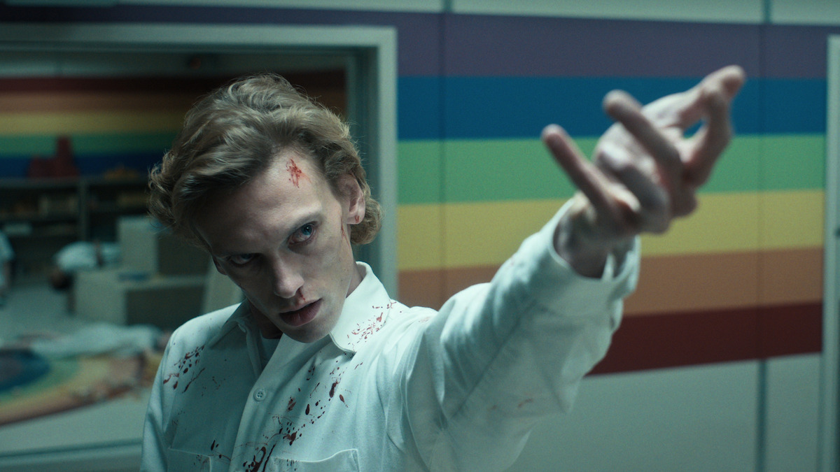 A pale man in a blood-splattered white shirt stands in a clinical, rainbow-striped room, extending his hand menacingly, creating a tense and eerie atmosphere.