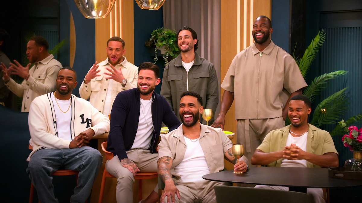Seven men sit and stand in a warmly lit room with plants and gold accents, smiling and laughing together in a relaxed, social atmosphere.