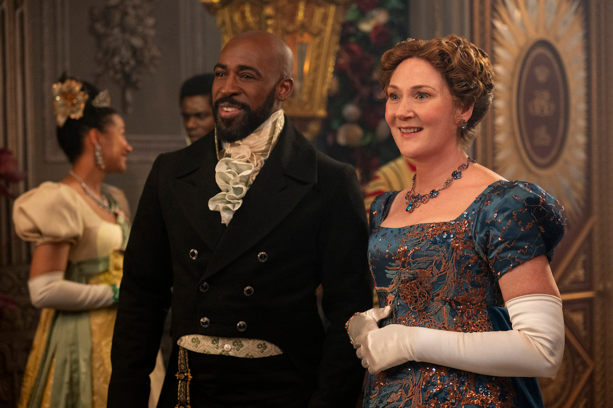 A man and woman in elegant Regency-era attire stand in a lavish ballroom, smiling and wearing formal gloves, with ornately decorated walls and other guests in the background.