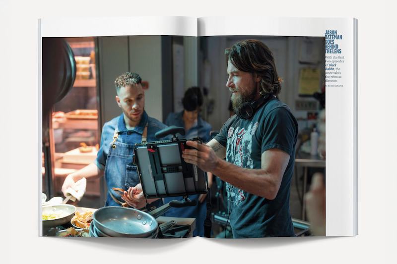 Two men in a kitchen, one cooking at a stove, the other holding film equipment, appearing to discuss a shot for filming. The environment is busy and professional, with kitchen tools and food visible on the counters.