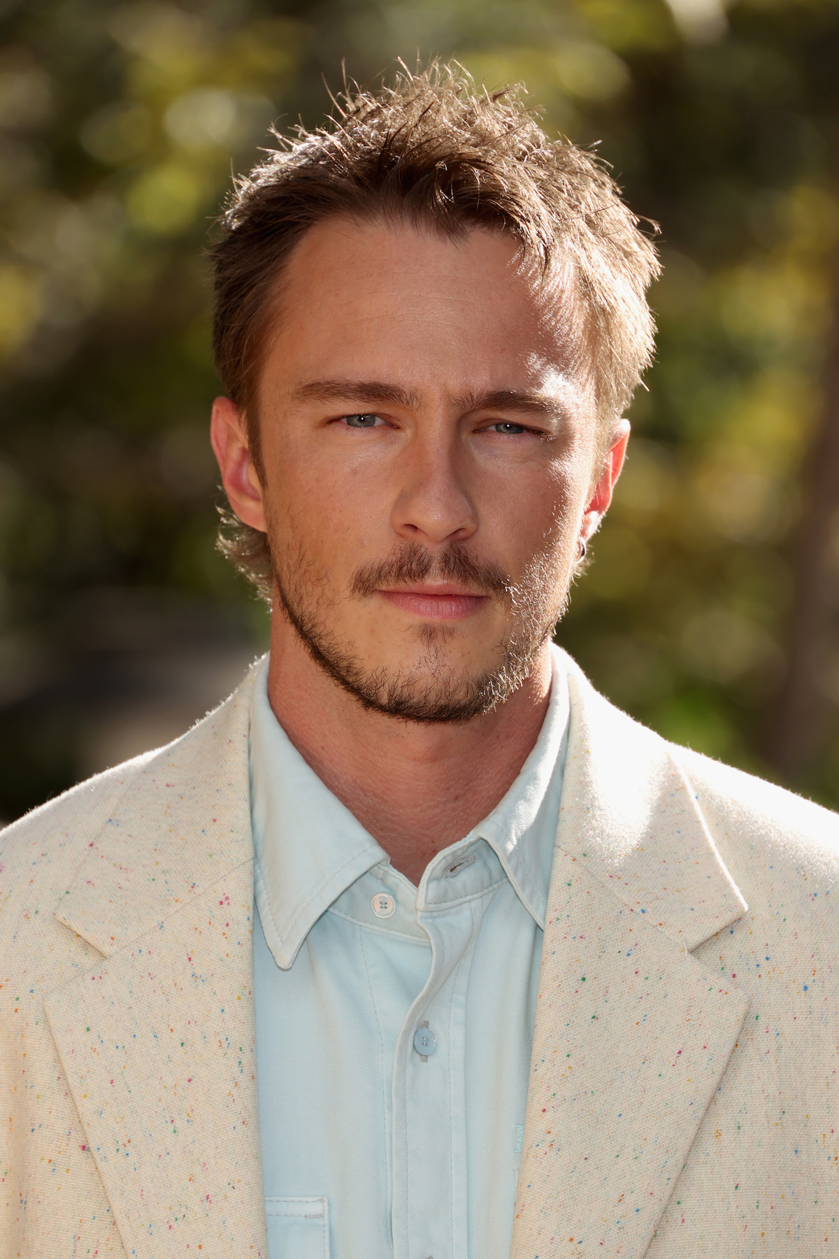 Drew Starkey with short brown hair and a trimmed beard wearing a light-colored jacket and blue shirt, standing outdoors with greenery and sunlight in the blurred background.