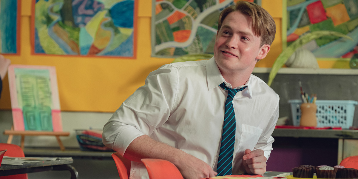A young man in a white shirt and striped tie sitting in a colorful classroom with artwork on the walls, looking off to the side and smiling, art supplies and paintings visible around him.