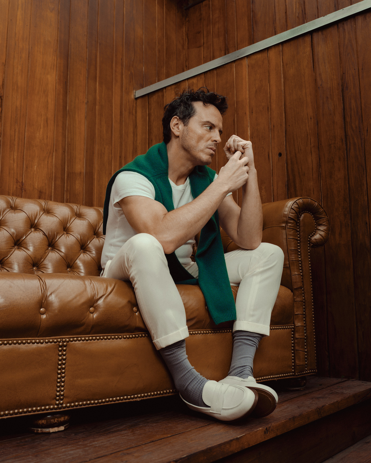 Andrew Scott wears white pants, shirt, and loafers, and a green sweater draped over his shoulders.