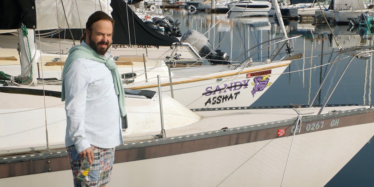 Man in preppy clothing stands on a marina dock holding a drink, with several sailboats moored in the background; one boat named "Sandy Asshat" is visible. Bright, sunny setting with calm water reflections.