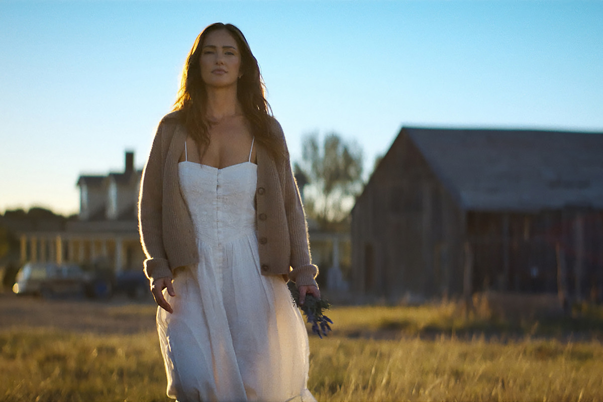 Woman in white dress and cardigan walks outdoors at sunset in a rural setting, holding flowers, with grass field, barn, and farmhouse in the background.