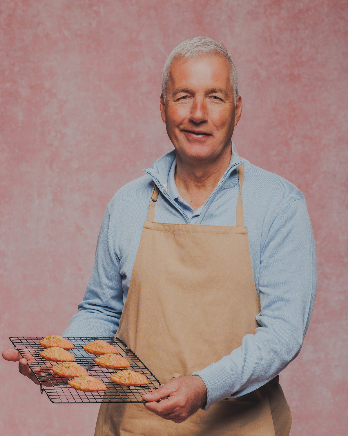 Older man wearing a beige apron, standing in front of a pink textured background, holding a cooling rack with freshly baked pastries, looking at the camera and smiling.