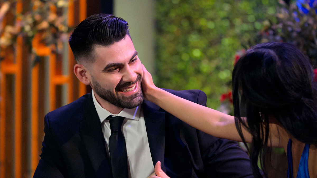 Mike Gibney in a suit smiling while Emma Betsinger gently touches his face, both seated in a warmly lit environment with greenery and floral decor in the background.