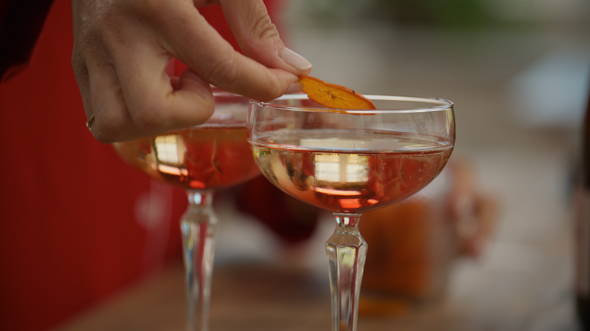 Hand garnishing a cocktail with a dried fruit slice in a coupe glass, two drinks on a wooden table, relaxed atmosphere, natural light, social setting.