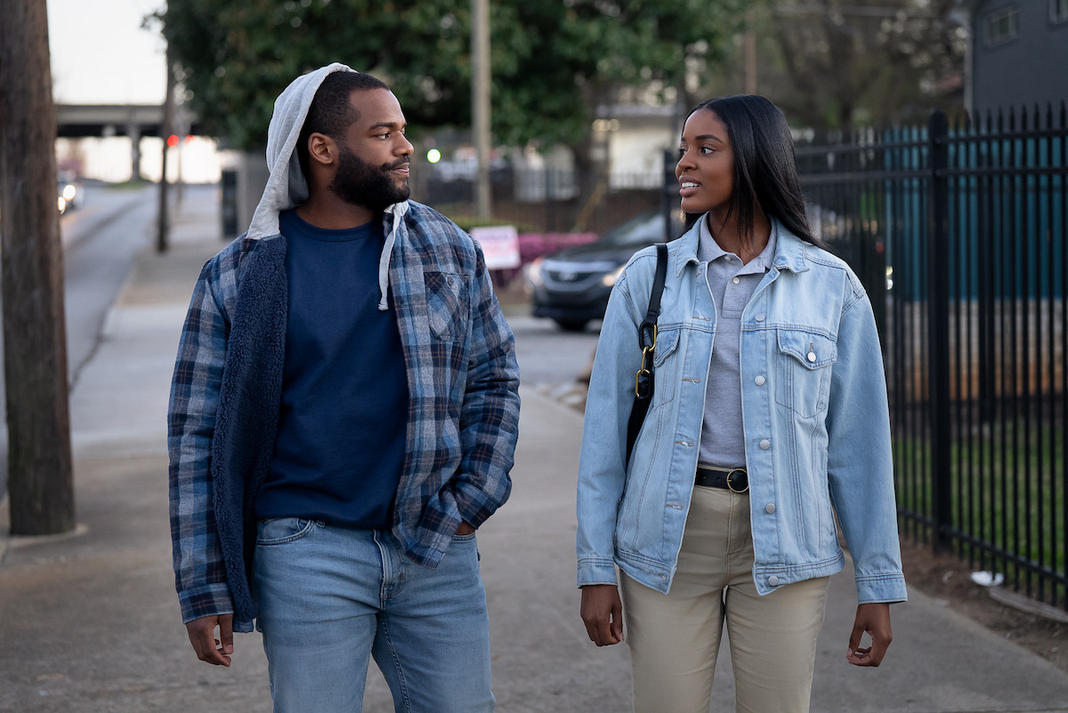 Two people walking on a city sidewalk, wearing casual jackets and jeans, talking and smiling in an urban outdoor setting with trees, cars, and fences visible in the background.