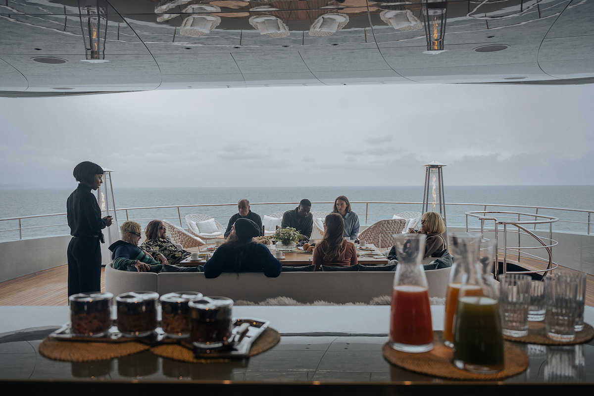 A group of people relax on a yacht deck overlooking the sea on a cloudy day, drinks and juice on a bar in the foreground, creating a calm, luxurious, and social atmosphere.
