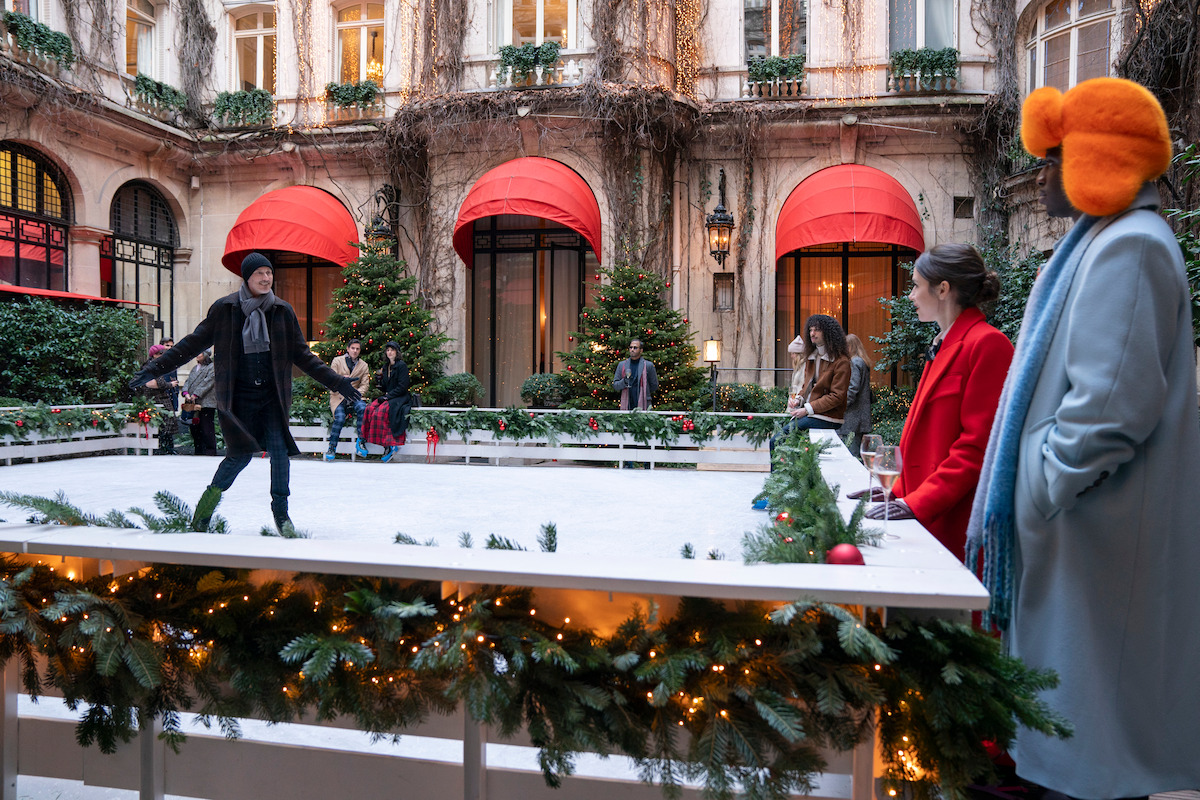 Bruno Gouery as Luc ice skates in a courtyard, as Lily Collins as Emily and Samuel Arnold as Julien watch him in Season 4 of ‘Emily in Paris’