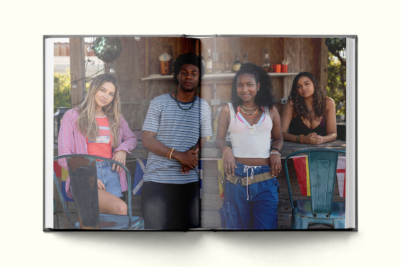 Four young people pose casually in an outdoor setting, sitting and standing in front of a rustic wooden structure decorated with hanging plants, giving a relaxed, summery vibe.