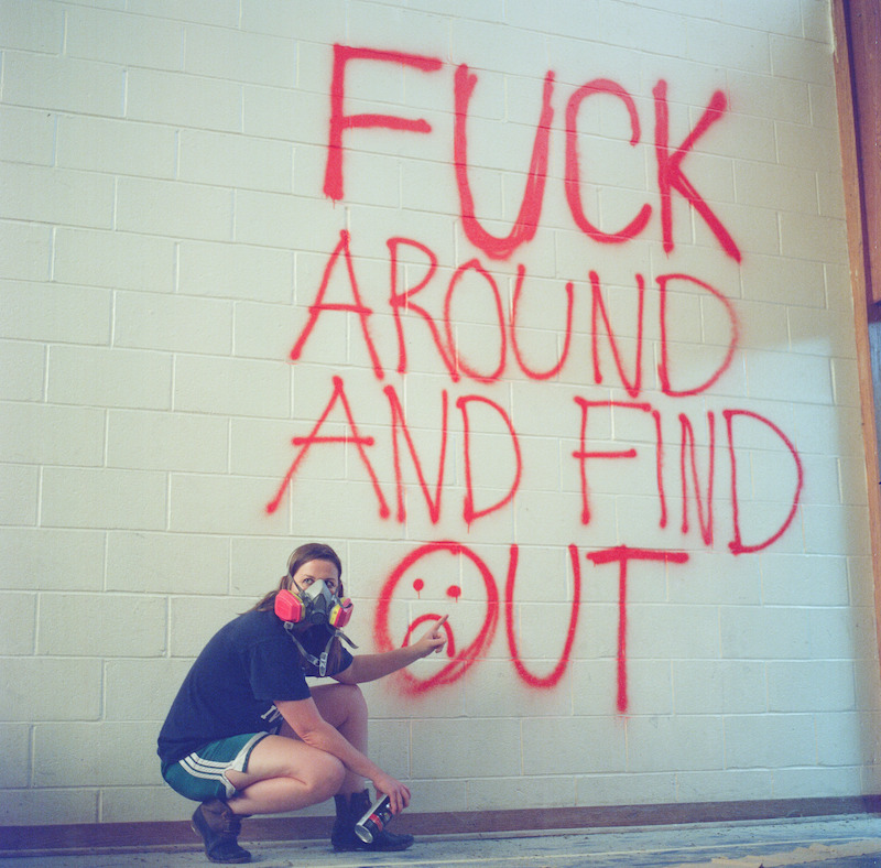 Katherine Kubler squatting in front of graffiti that reads "Fuck around and find out."
