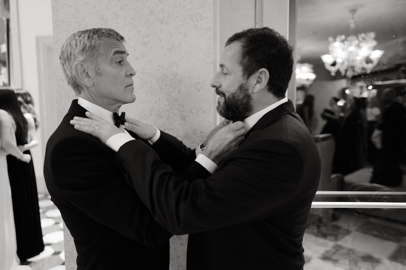 George Clooney and Adam Sandler adjust each other's bow ties in a hotel while wearing tuxedos. 