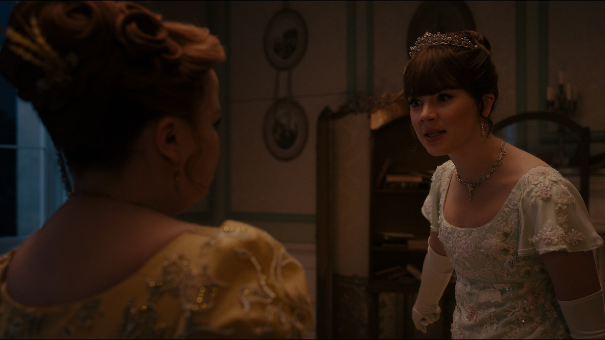 Two women in elegant 19th-century dresses talk intensely in a dimly lit, ornately decorated room with framed art on the walls, one wearing a tiara and gloves, suggesting a historical or period drama setting.