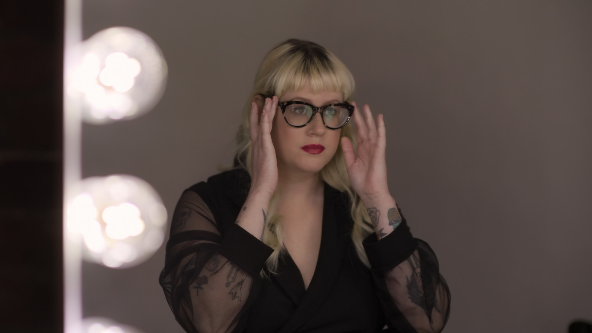 Person with blond hair and glasses adjusts frames while sitting at a makeup vanity with lit bulbs, wearing a dark sheer-sleeved blouse; neutral background.