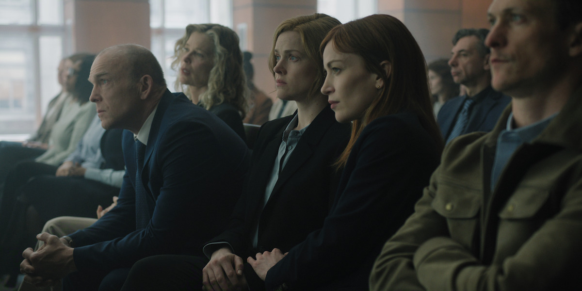 People sit in rows in a courtroom or formal setting, dressed in business attire, attentively focused on proceedings taking place out of frame, with large windows and neutral lighting in the background.