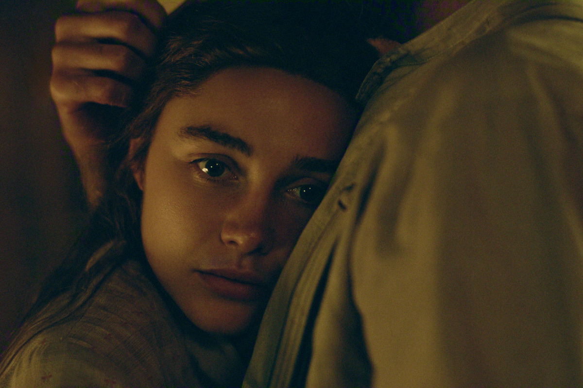 A young woman with expressive eyes rests her head against a man's chest in a dimly lit, intimate setting, conveying a sense of comfort and emotion.