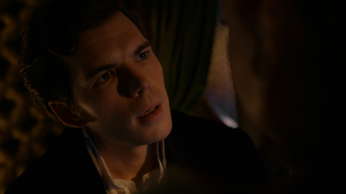 A man in period clothing sits in a dimly lit room with patterned walls and green drapery, looking intently at someone just out of focus in the foreground. Warm, soft lighting highlights his thoughtful expression.