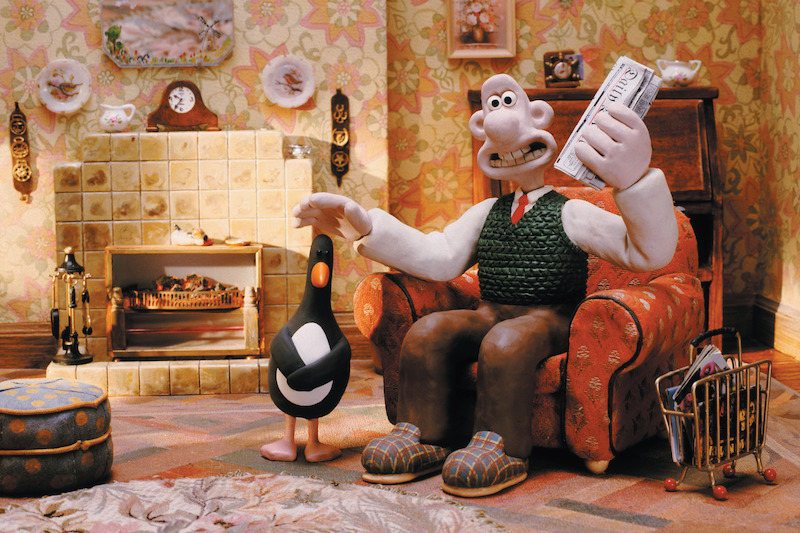 Wallace wears brown pants, a white shirt, and green vest in his living room.