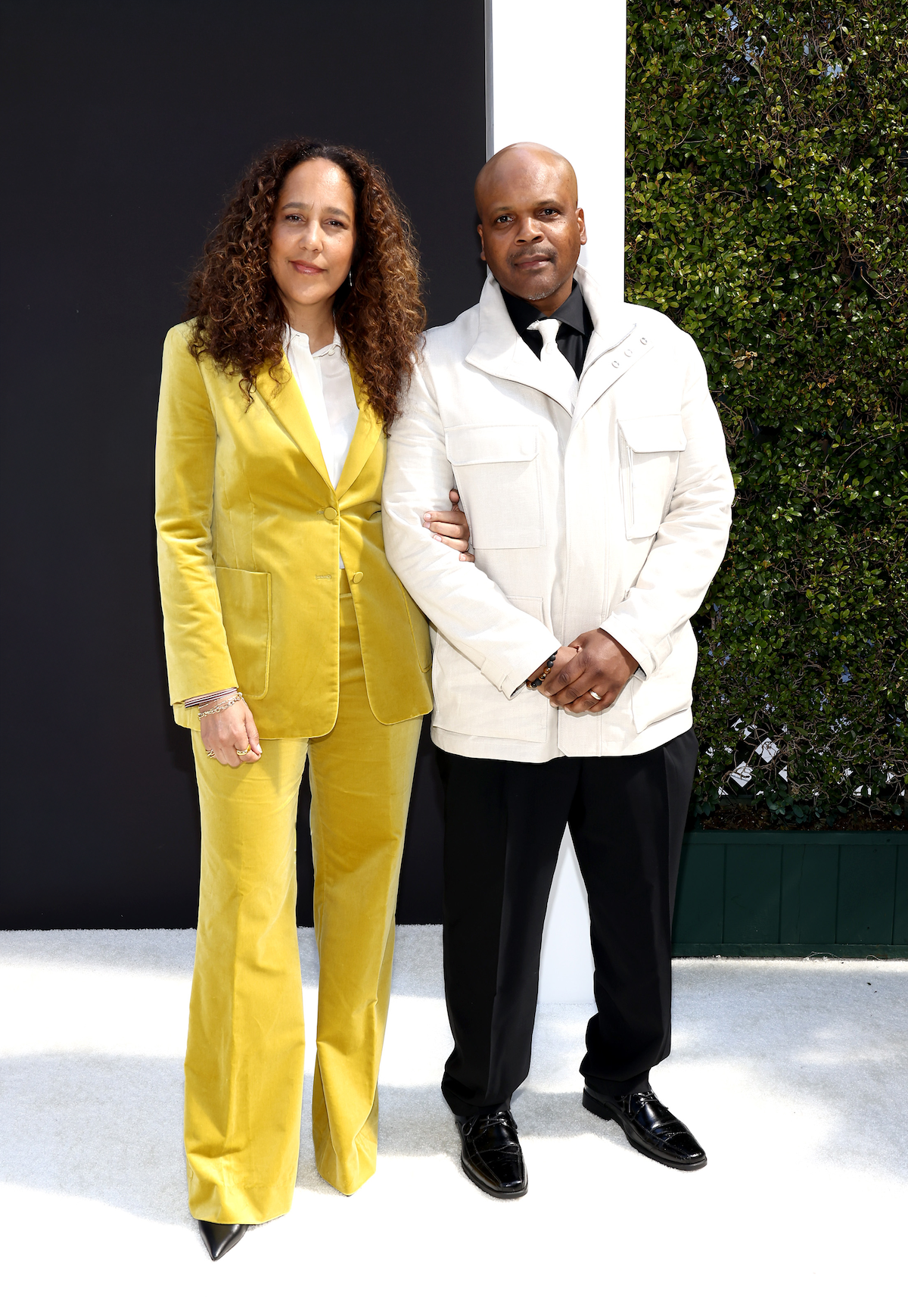 Two people pose together outdoors against a modern, stylish backdrop. The woman wears a yellow suit and white blouse, while the man wears a white jacket with black pants and shoes. The setting appears formal and well-lit.