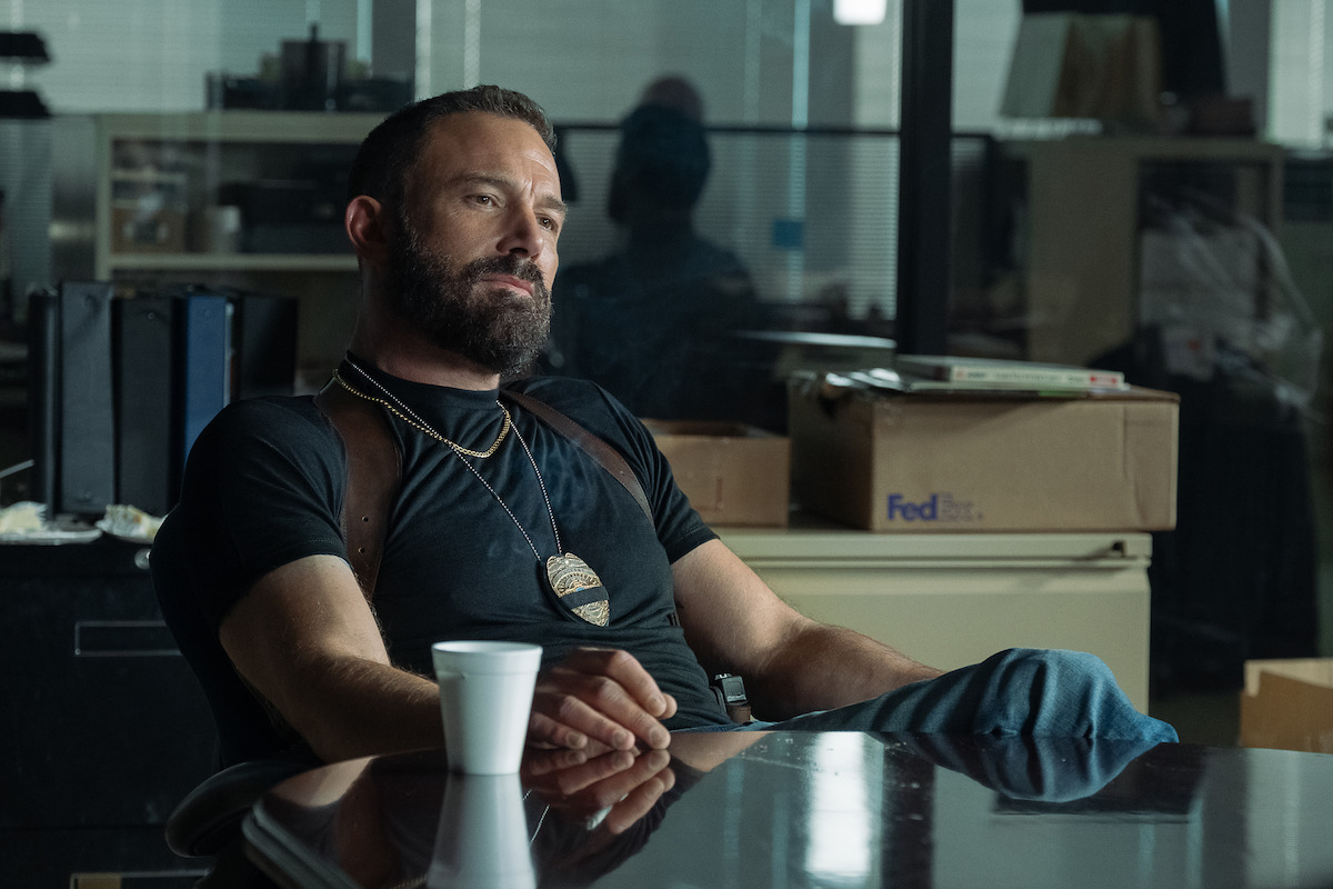 Bearded man wearing a badge and shoulder holster sits relaxed at a desk in an office, coffee cup in hand, with boxes and files in the background, giving a detective or law enforcement vibe.