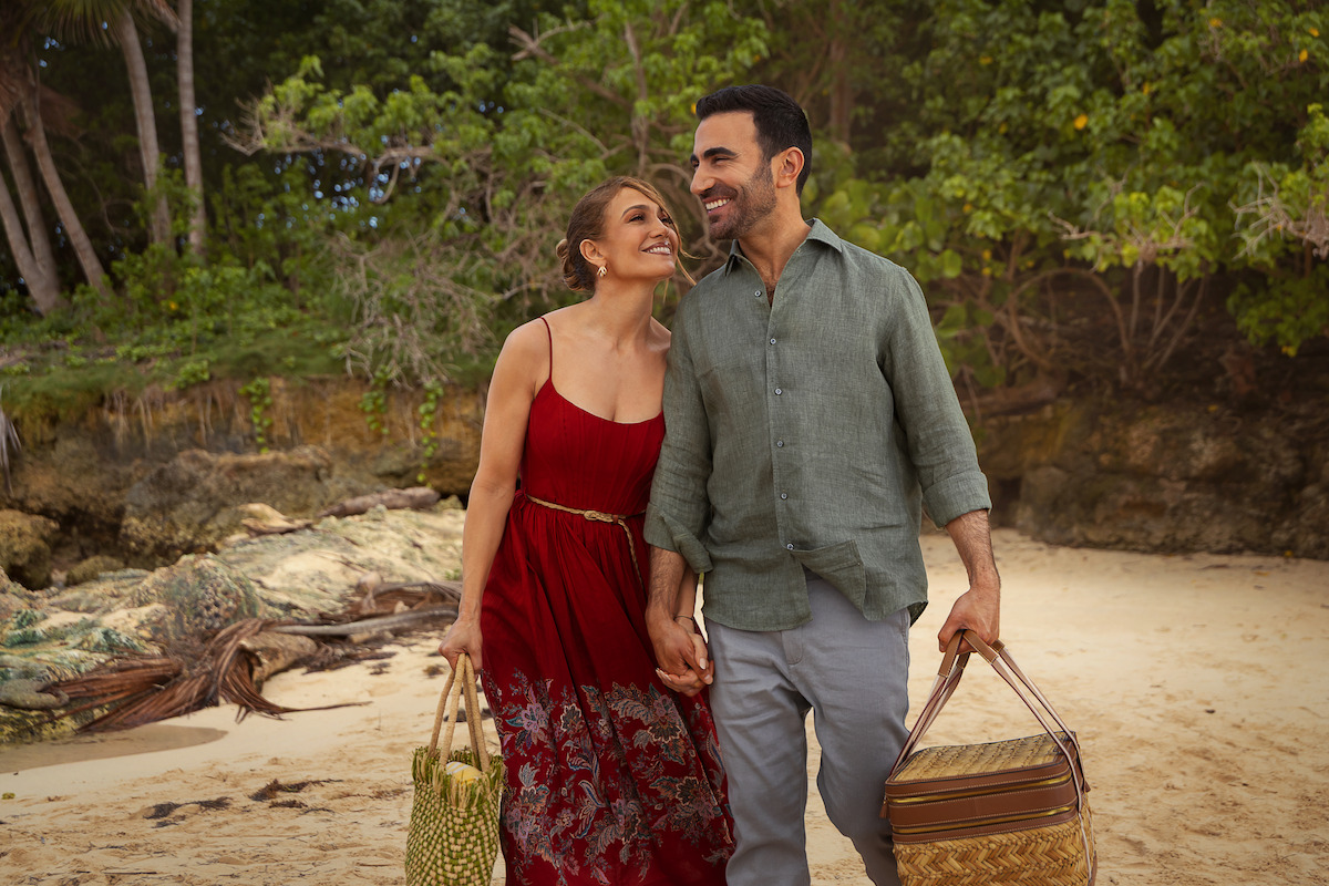 A smiling couple walks hand in hand on a sandy beach, surrounded by tropical trees, each carrying picnic baskets, enjoying a sunny, relaxing day outdoors.
