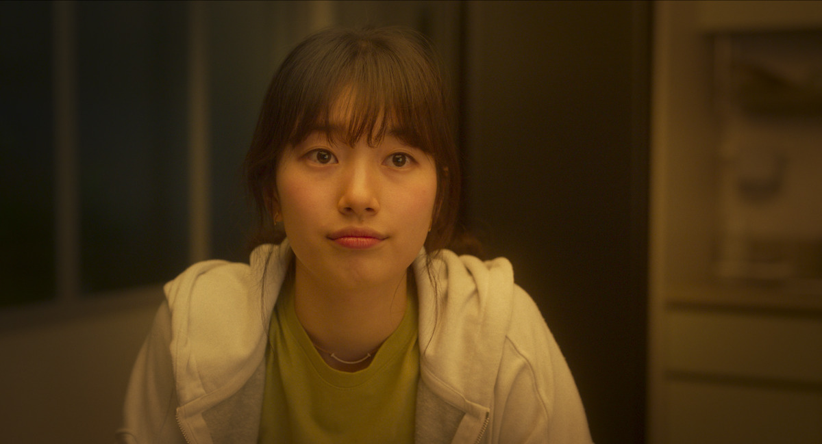 Suzy as Gu Jung-in sits in a warmly lit room at night while wearing a white hooded sweatshirt in an image from the film ‘Wonderland.’