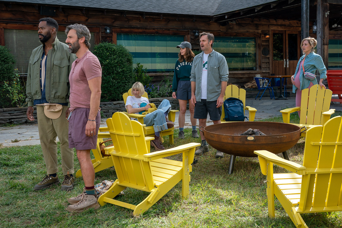 Six adults outdoors in front of a wooden cabin, standing or sitting around a fire pit with yellow Adirondack chairs on grass, dressed in casual summer or hiking clothes, suggesting a group camping or retreat setting.