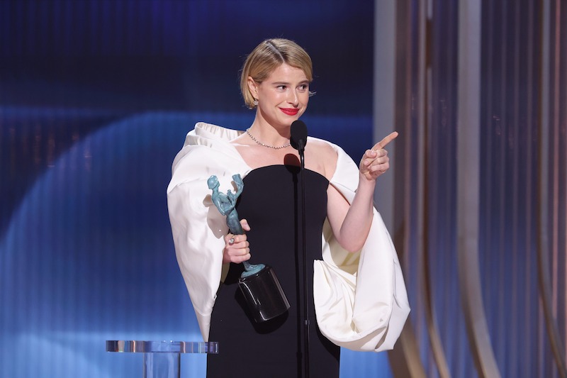 Jessie Buckley in elegant dress holding a trophy, speaking at a microphone on stage with dramatic lighting and vertical gold and blue background, likely at an awards ceremony.