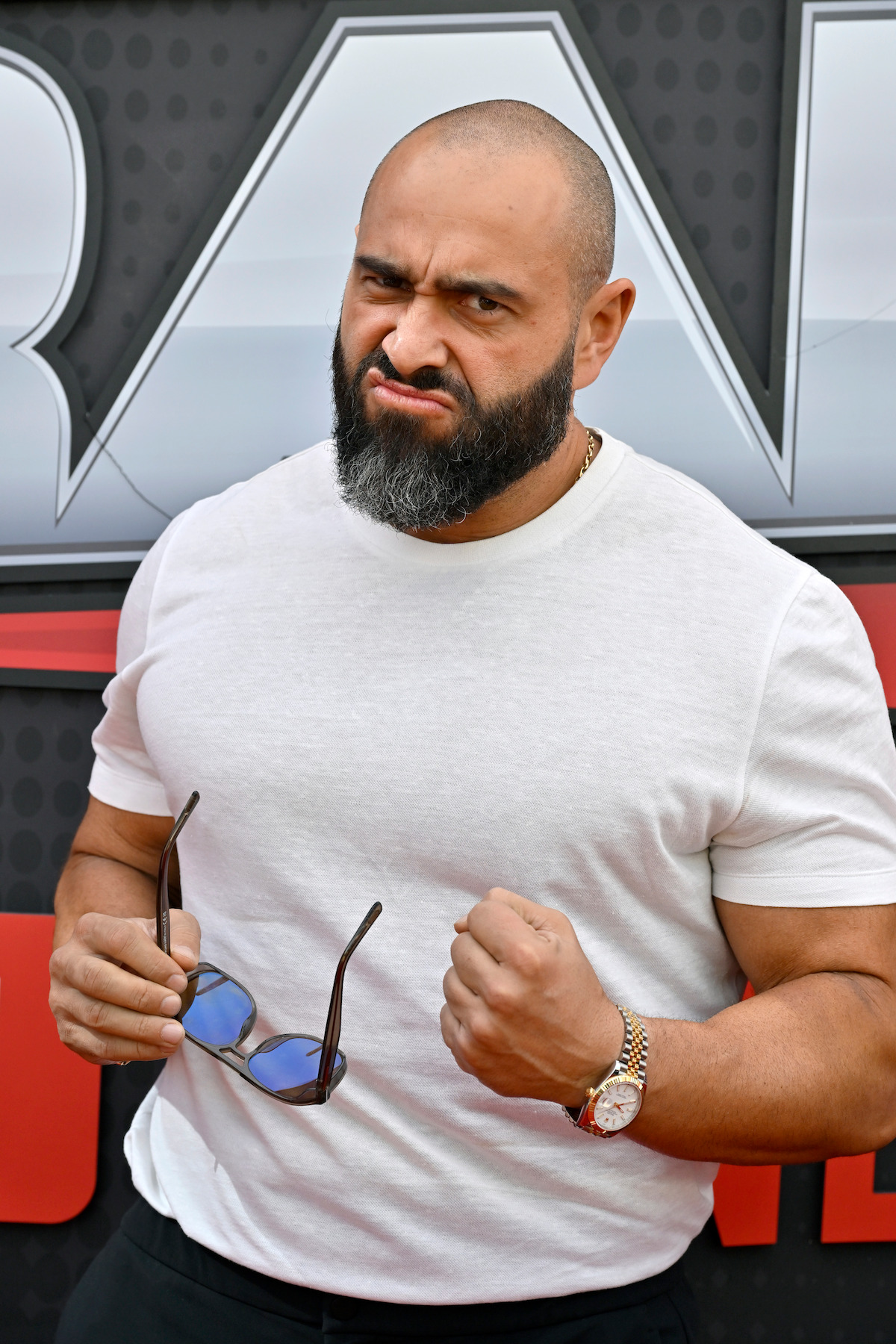 Bearded man in a white t-shirt holding sunglasses and making a fist, standing in front of a graphic background at an event, posing with a serious expression.